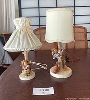 Two Hummel figurine table lamps on a wooden table showing fabric shades and detailed bases with small painted children figures.