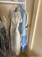 Photo of four vintage-style clothes hung in closet including wedding dress, blue skirt suit, beige skirt and top, and varsity sweater