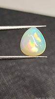 Pear-shaped cabochon Ethiopian opal gemstone held by tweezers showing colorful internal flashes against a dark background.