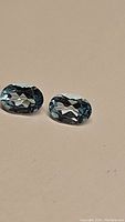 Close-up photo of two oval cut, sky blue topaz gemstones placed on a flat surface showing color and faceting details.