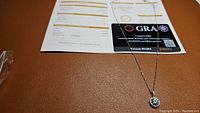 Photo of the 1 carat certified moissanite pendant necklace on a brown surface with certificate documents visible