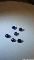 Photo showing six small round blue sapphire gemstones arranged randomly on a plain light surface, displaying their deep blue colors and round faceted cuts.