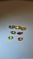 Seven small polished sapphire gemstones in mixed colors of green, yellow, orange and red laid out on white surface