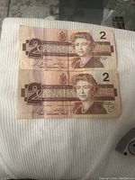 Top front view of two Canadian two-dollar bills from 1986, showing Queen Elizabeth II portrait and denomination markings, placed flat on a white ribbed surface.