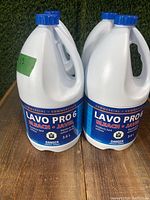 Four white plastic bottles with blue caps of Lavo Pro6 commercial bleach, arranged outdoors on grass.
