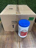 Closed cardboard box and one tube of BioSan Wipes with blue lid placed in front, on wooden surface with grass background.