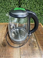 Full clear glass kettle with black handle and lid, standing on wooden surface with green background.