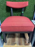 Front view of the chair showing padded red fabric seat and dark wooden legs on wooden floor against green grass-like wall