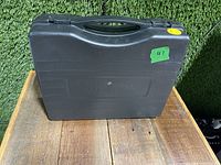 Black plastic carrying case for a portable gas stove placed on a wooden surface against greenery.
