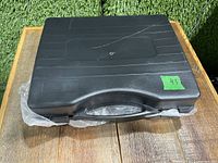 Black plastic carrying case with handle containing the portable stove. Green lot number sticker visible.