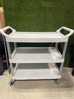 Front view of the white Rubbermaid utility cart with three shelves and handles on both sides, placed against a green backdrop.