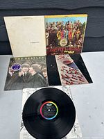 Five Beatles vinyl LP records displayed with original album covers showing on surface, featuring iconic designs including 'White Album', 'Sgt. Pepper', 'Meet The Beatles!', 'Abbey Road', and 'Rubber Soul'.