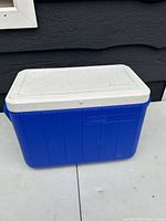 Blue rectangular Coleman cooler with white lid placed against a dark background.