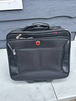 Black SwissGear laptop briefcase standing upright on pavement with telescoping handle retracted, displaying front zippered compartments and red SwissGear logo.
