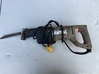 Electric reciprocating saw placed horizontally with visible power cord wrapped around it, showing blade, trigger handle, and metal body.