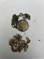 Closed view of one pocket watch with floral engraving and attached chain, with a gold-tone necklace/chain below.