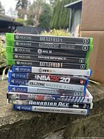 Stack of 11 video game cases with visible titles for Xbox One and PS4, photographed outside on a step.