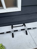 Silver and black pull up bar with padded grips laid flat on white table against blueish wall. Shows the entire bar and its multiple grip locations.