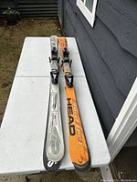 Photo showing two skis, one silver and one orange, both with bindings and marked Head, placed on an outdoor bench against a dark gray wall.