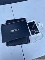 Photo of a black Sony Vaio laptop closed and placed on top of a white tablet device with a charger cable connected, displayed on white surface.