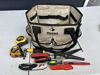 Husky tool bag open with various tools laid out including tape measures, pruning shears, screwdrivers, box cutter and metal clamp.