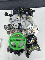 Photo showing a variety of wired video game controllers and a green Nintendo GameCube steering wheel accessory placed on a light colored surface.