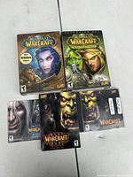 Photo showing five World of Warcraft game boxes and manuals on a white surface, including original packaging and manuals for base game and expansion sets.