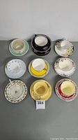 Top-down view of 10 assorted cup and saucer sets showing different patterns and colors including turquoise, black floral, white with blue flowers, yellow, multicolor floral, cream with sprig, red and yellow sets.
