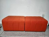Two orange fabric modular ottomans shown side by side from a front angle on a floor against a wall.