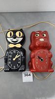Front view of both clocks side by side showing black Kit Cat clock with yellow eyes and bow tie, and red Klocker Spaniel dog clock.