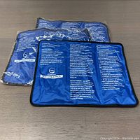 Three blue rectangular AllSettHealth hot/cold therapy packs lying on a wooden surface, with printed therapy instructions visible on the top side of each pack.