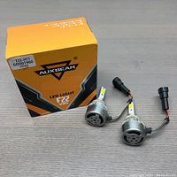 Pair of Auxbeam F22 Series LED lights with their original orange and black packaging box, shown on a wood surface. Each light has a vented aluminum housing with a rear fan and attached H11 connector cable.