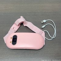 Pink cat eye mask warmer and massager with adjustable strap laid flat on a gray surface with a USB charging cable beside it.