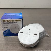Photo of the CO/Smoke Alarm device next to its original box, showing front design and packaging.
