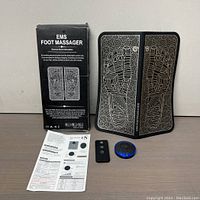 Front view of EMS Foot Massager box, unfolded massager pad with foot pattern, remote control, circular blue controller, and user manual laid flat on table.
