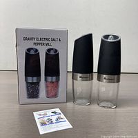 Two Gravity Electric Salt and Pepper Mills with transparent bottom chambers showing the peppercorns and pink salt, black ergonomic tops, brand logo silver bands, instruction sheet, and product box.