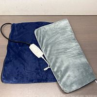 Large blue heating pad with grey removable cover flat on table with white control attached by black cord.