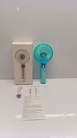 Mini handheld teal fan alongside its box, USB charging cable, and instruction sheet.