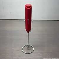Front view of the red handheld milk frother standing upright in its chrome circular stand on a gray surface.
