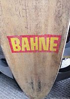 Close-up of colorful Bahne brand logo on the wooden bamboo longboard deck showing wear and scratches.