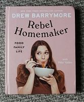 Front cover of Rebel Homemaker cookbook by Drew Barrymore showing pink cover and photo of Drew eating noodles.