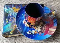 Blue 50 years anniversary plate with Donald Duck and colorful Disney mug on top, small clear glass beside them, and photo album with Tinkerbell cover underneath.