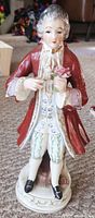 Front view of porcelain colonial man figurine showing detailed facial features, red coat, white pants, and flower in hand.