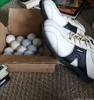 Photo showing golf cleats and box of golf balls on carpeted floor.