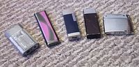 Five assorted vintage lighters displayed in a row on carpet; varying colors and wear.