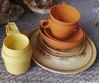Stack of Melmac dishes with floral design and orange Melmac cup and saucer on top, alongside nested yellow Tupperware measuring cups.