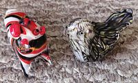 Two glass paperweights on a gray textured surface; one bird-shaped with swirling black, white, and purple glass, the other a striped tiger/dragon figure with red, black, white, and yellow.