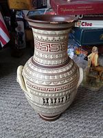 Front and side view of Greek geometric style vase showing intricate red and black patterning with stylized figures and meander design. Cream base color and brown rim.