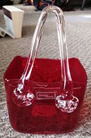 Photo showing red crackle glass vase shaped like a small handbag with clear glass handles sitting on carpet.