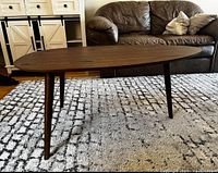 Side view of walnut coffee table with three tapered legs on a patterned rug, with leather sofa and cabinet in background.